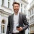 Professional in a gray suit and white shirt smiling confidently while holding a smartphone, standing in front of classical white buildings with arched windows and elegant architectural details.