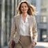 Professional woman in a beige business suit and white blouse walking confidently in an urban environment, with wind-swept hair and carrying a leather portfolio, against a blurred city background.